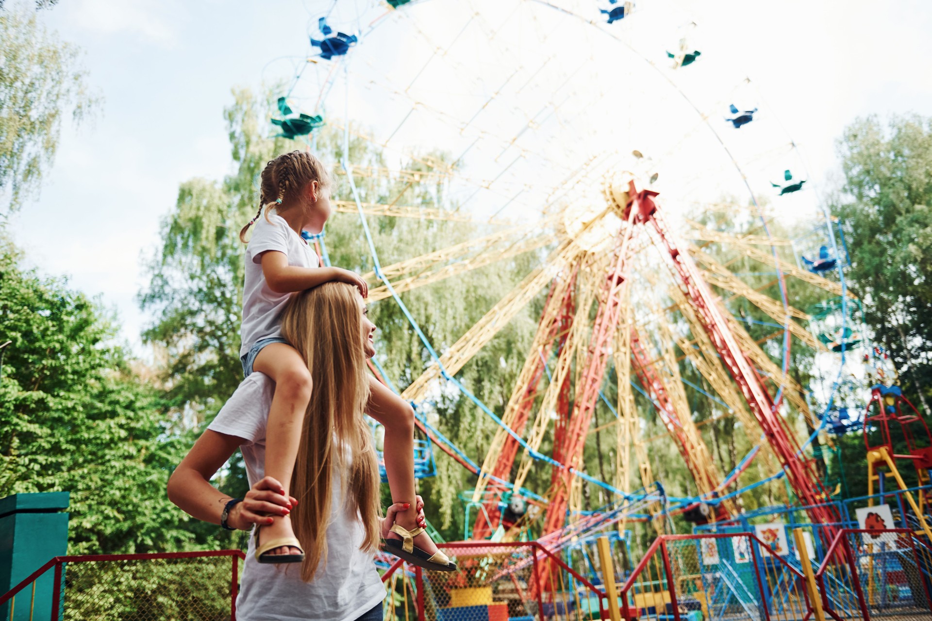 Daughter sits on the shoulders. Cheerful little girl her mother have a good time in the park together near attractions